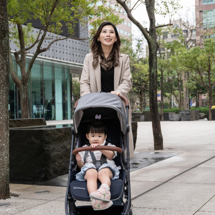 “Woman pushing the UPPAbaby MINU V3 stroller in Greysen with a toddler seated, walking through a tree-lined urban plaza.”