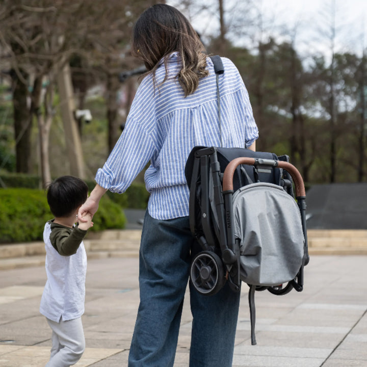Woman carrying the folded UPPAbaby MINU V3 stroller in Greysen on her shoulder while holding a child’s hand outdoors.