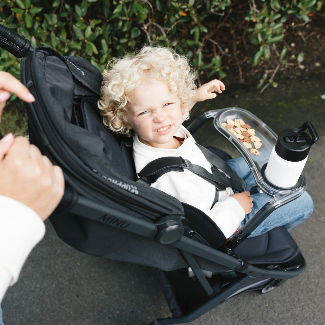 Child seated in UPPAbaby Minu V3 stroller using snack tray with cup holder and snacks, outdoor setting