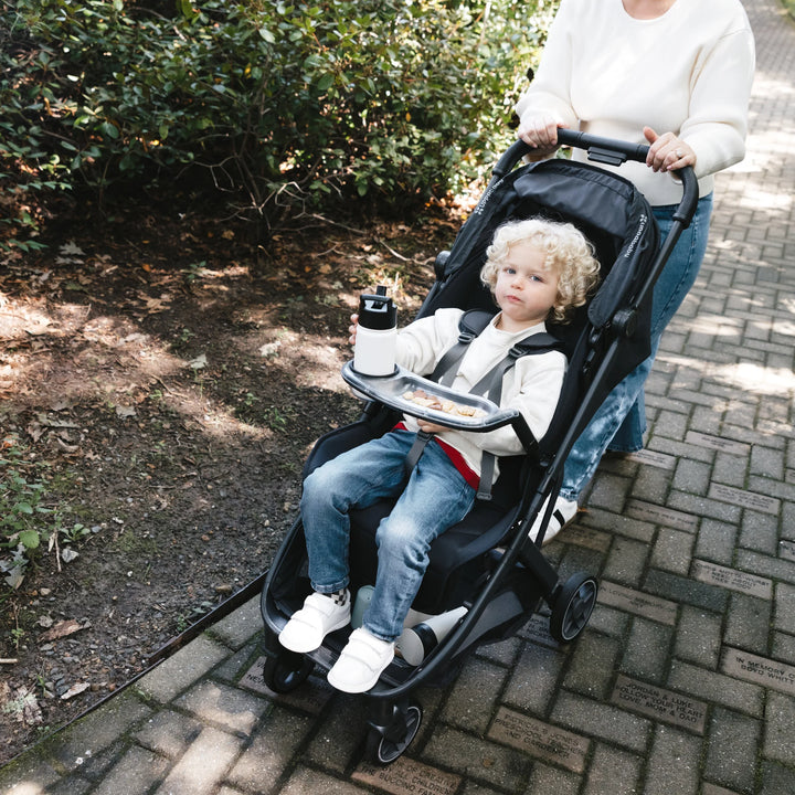 Child seated in UPPAbaby Minu V3 stroller using snack tray with cup holder and drink, pushed along outdoor path