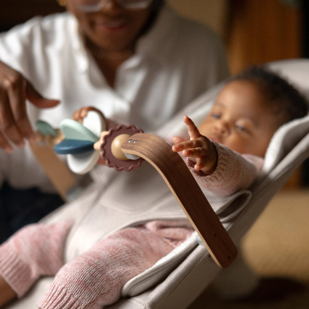 Close-up of a baby reaching for the UPPAbaby Mira Wonder Wheel wooden toy bar while seated in the UPPAbaby Mira bouncer, with a parent spinning the sensory toys in a warm home environment.