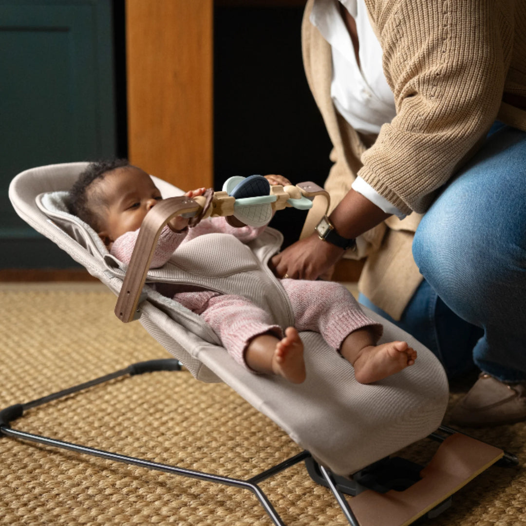 Baby relaxing in the UPPAbaby Mira bouncer while engaging with the UPPAbaby Mira Wonder Wheel wooden toy bar, as a parent adjusts the toy bar in a cosy home setting.