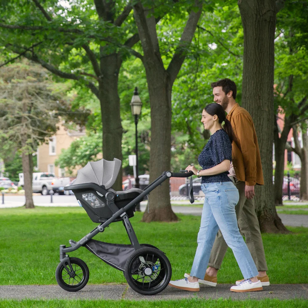 Two happy parents are pushing their baby in the Mesa i-Size infant car seat on the Ridge chassis through a green park area.