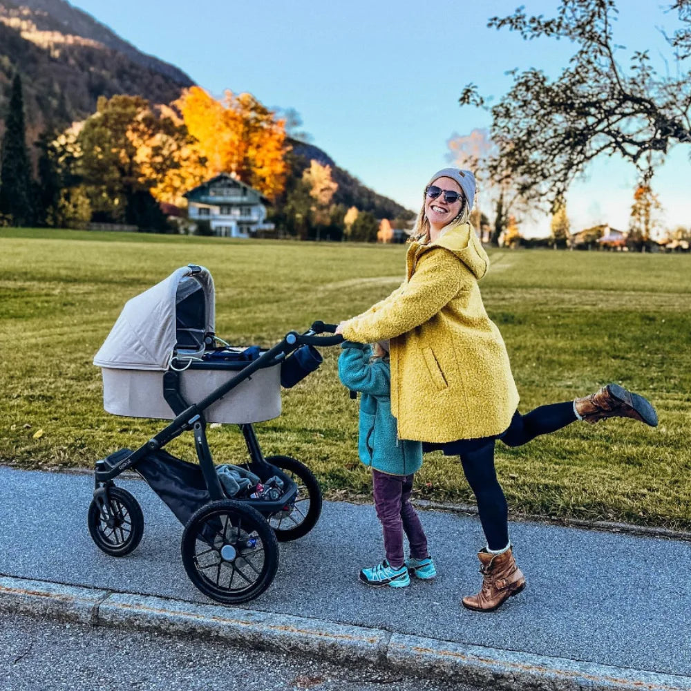 This image shows a happy parent pushing the Ridge stroller with the help of their child while the baby is in the carrycot accessory.