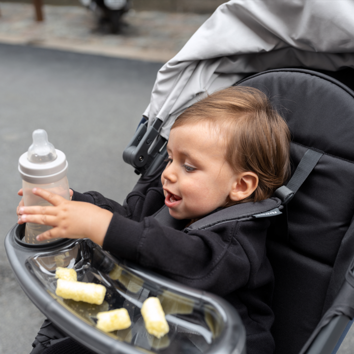 This is a lifestyle image of the Ridge snack tray on the stroller with a toddler placing his sippy cup back in the cup holder slot and there are a few snacks in the recessed dish part