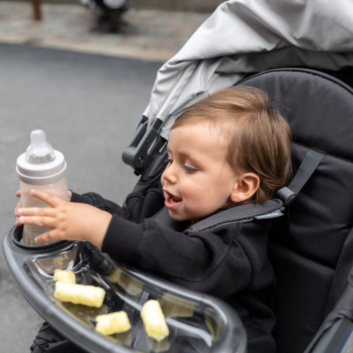 Toddler using the UPPAbaby RIDGE stroller snack tray with cup holder, holding a bottle and snacks while riding in the stroller.