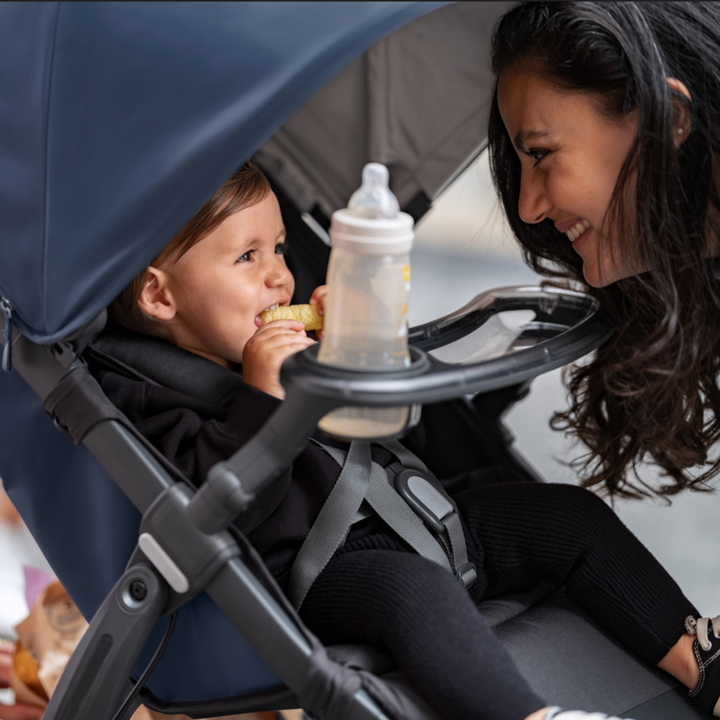 This is a lifestyle image of the snack tray in use on the Ridge stroller holding a baby bottle. The child is grinning and finishing a snack while the parent is smiling at the child.