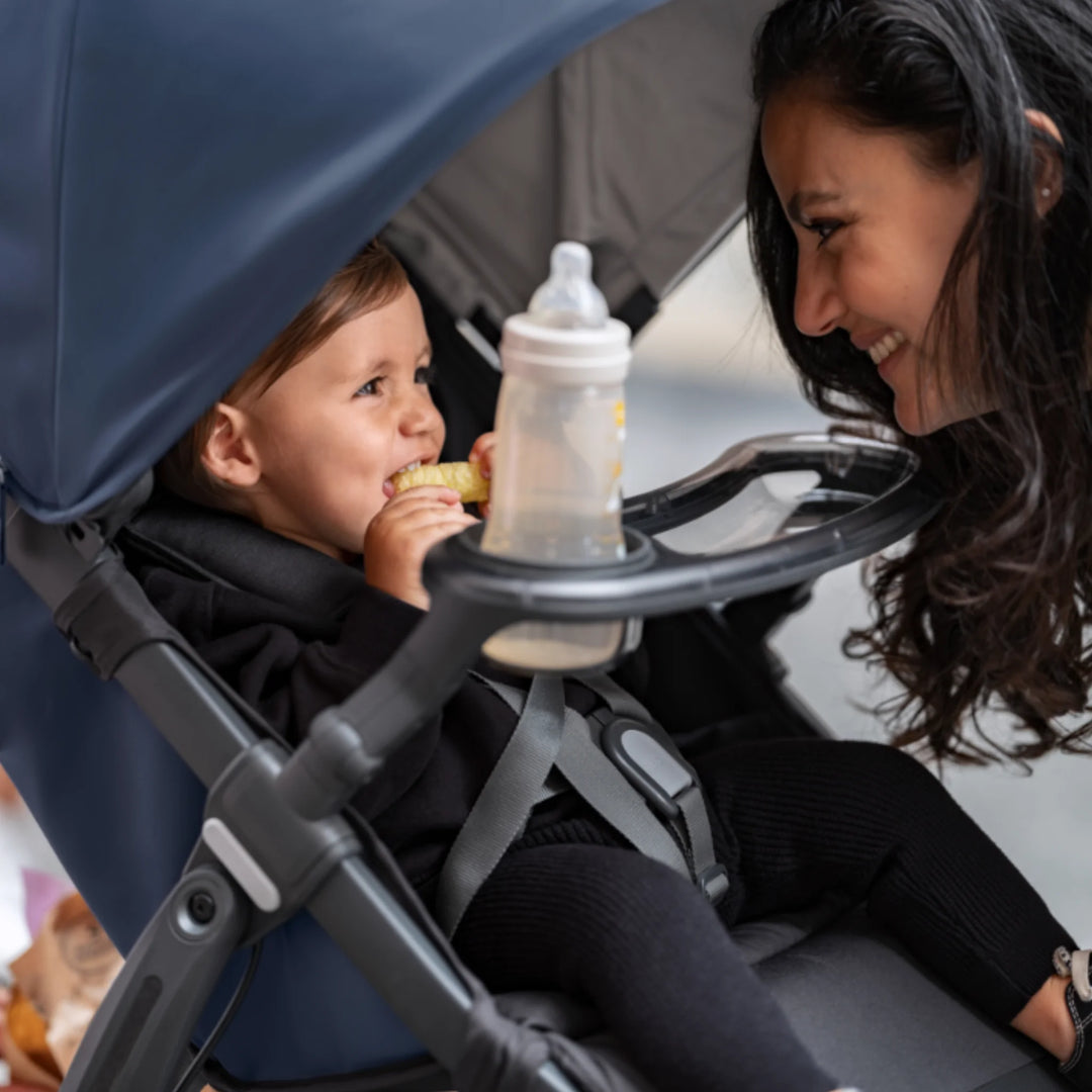 Baby enjoying snacks in the UPPAbaby RIDGE stroller snack tray with cup holder, while parent smiles beside the stroller.