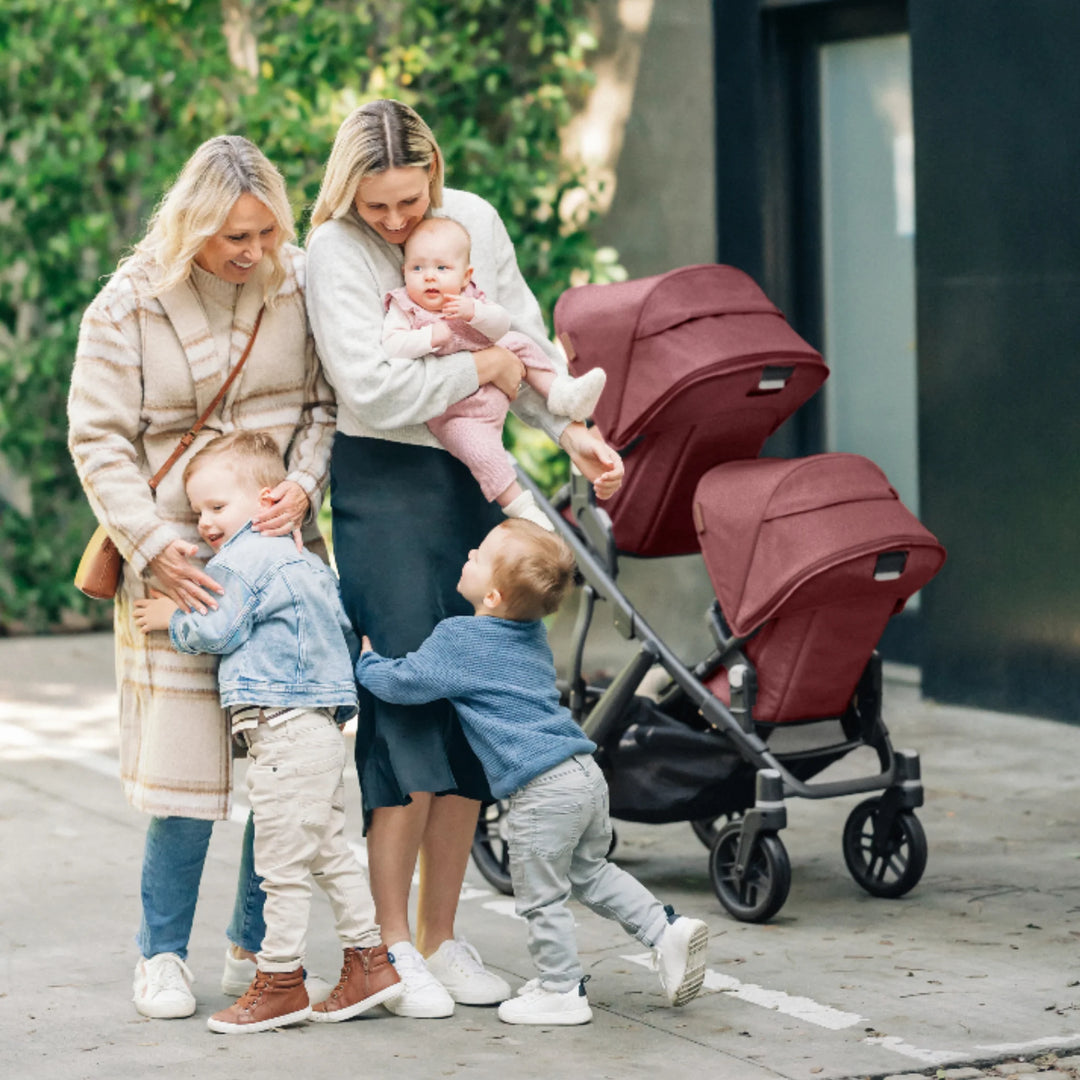 Family with two women and children beside an UPPAbaby Vista double stroller with Lucy red canopies in an outdoor lifestyle scene.