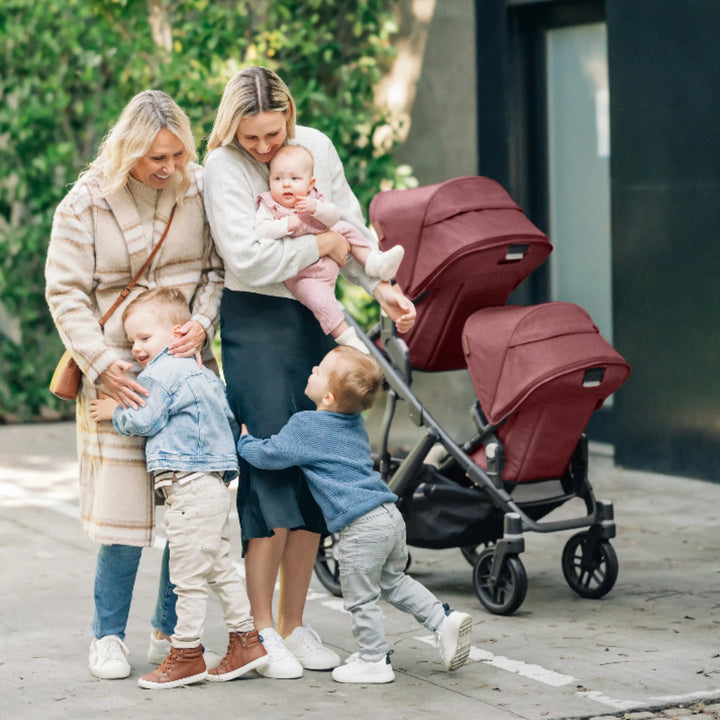 Family with two women and children beside an UPPAbaby Vista double stroller with Lucy red canopies in an outdoor lifestyle scene.
