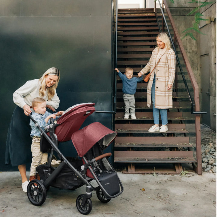 Two women with toddlers using an UPPAbaby Vista stroller configured as a double with Lucy red canopies outdoors by stairs.