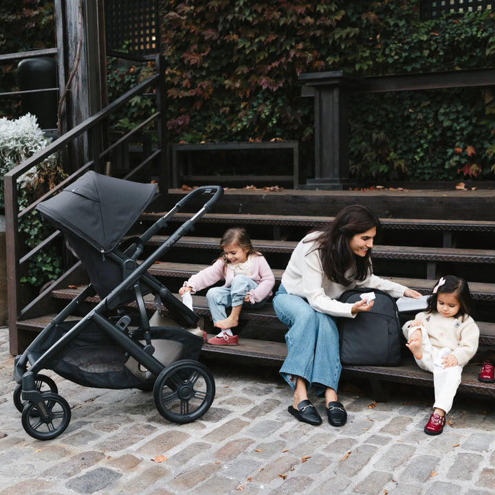 Mother sitting with two young children outdoors beside the UPPAbaby Vista V3 Jake stroller, showcasing family use of the black premium stroller during a relaxed outdoor moment on wooden steps.