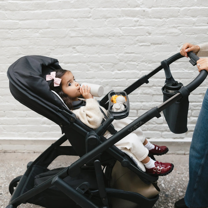 UPPAbaby Vista V3 Jake stroller in use, showing a toddler drinking from a bottle in the forward-facing seat while a parent pushes the stroller with the cup holder attached during an outdoor walk.