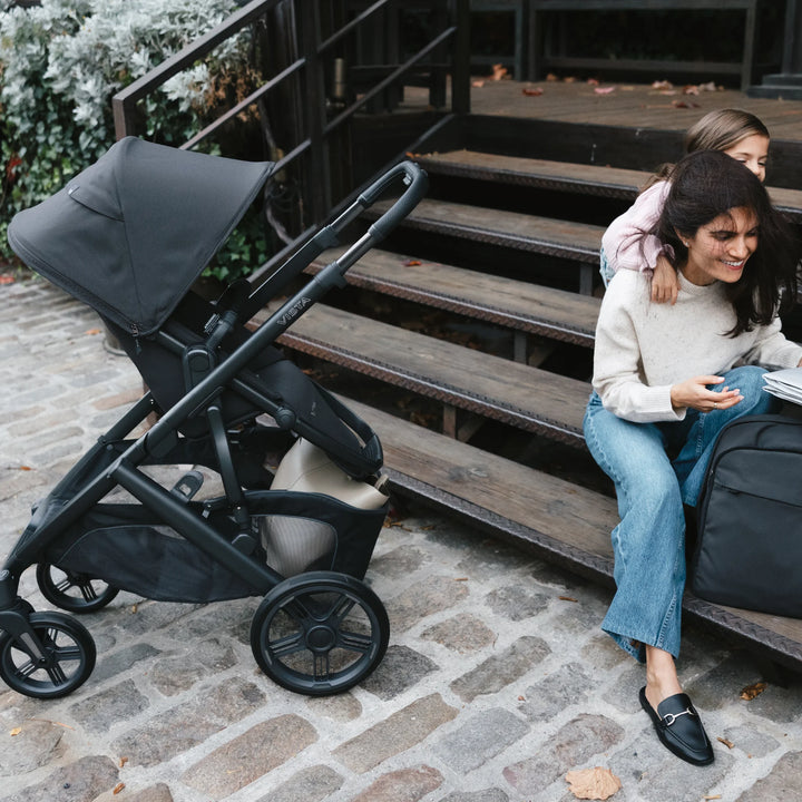 UPPAbaby Vista V3 Jake stroller in use outdoors with a mother sitting on wooden steps while her child hugs her from behind, showcasing the black premium stroller’s everyday family versatility and modern design.