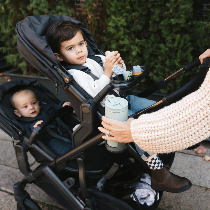 UPPAbaby Vista V3 Jake double stroller in use, showing toddler in the main seat and infant in the RumbleSeat while a parent hands a cup from the handlebar cup holder during an outdoor walk.