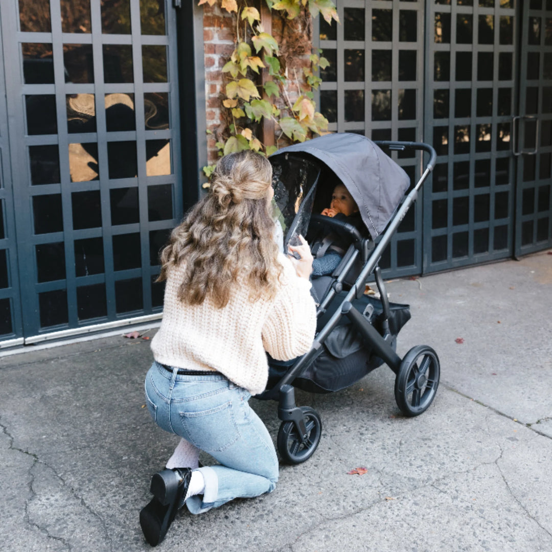 Parent kneeling to open and adjust the UPPAbaby Performance Rain Shield on a Vista V3 stroller, keeping baby protected from rain and wind.