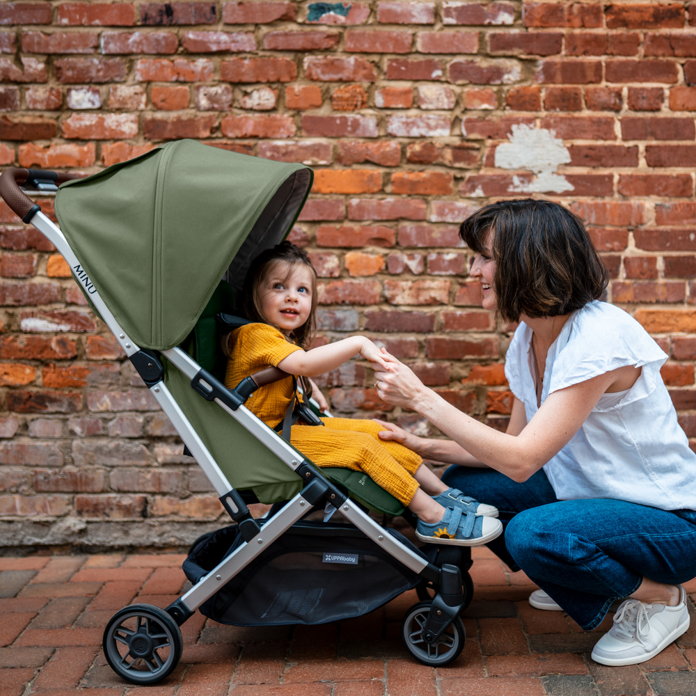 UPPAbaby Minu V2 lightweight travel stroller in Emelia which is a green fabric with chestnut REACH certified leather accents on a silver frame. This image shows the Minu in front of a red brick wall and a mother is kneeling down chatting to her toddler who is dressed in a bright yellow jumpsuit.