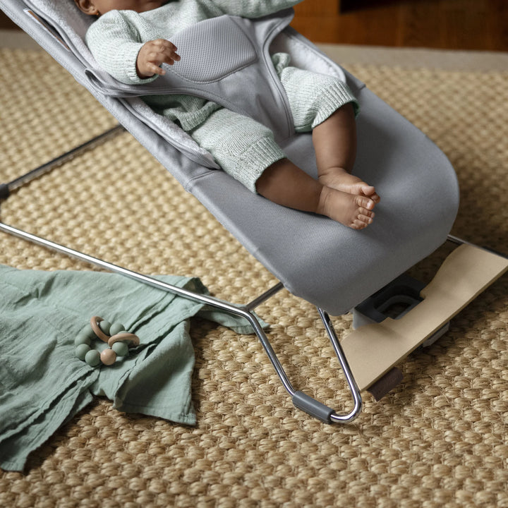 Close-up of a baby seated in the UPPAbaby Mira Bouncer in Stella grey, showing the ergonomic harness, breathable seat fabric, and sturdy lightweight frame with natural wood base on a textured woven rug