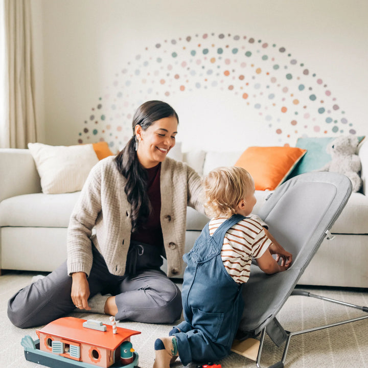 “Parent and toddler playing beside the UPPAbaby Mira Bouncer in Stella grey, highlighting the modern minimalist baby bouncer design with breathable mesh seat and sturdy steel frame in a bright, family-friendly living room.”