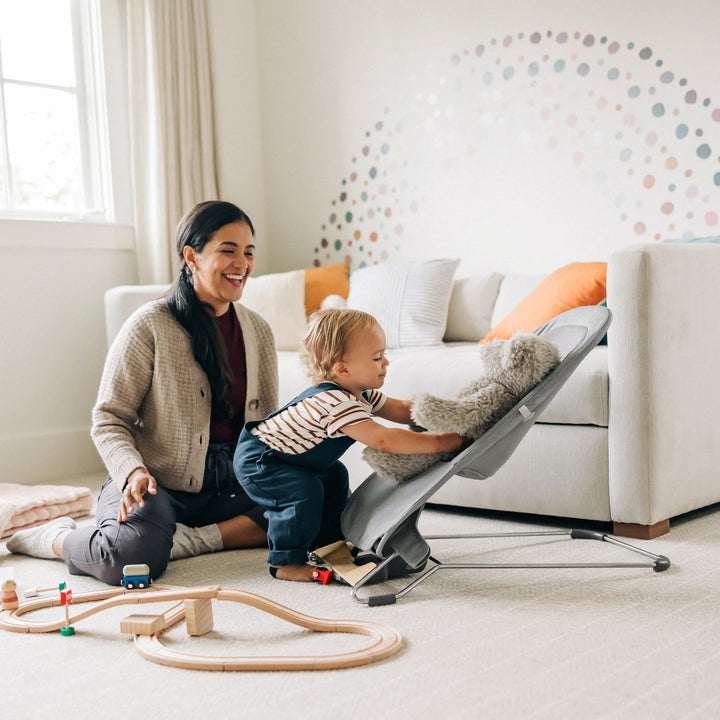 Parent and toddler playing in a bright family living room with the UPPAbaby Mira Bouncer in Stella grey, highlighting the modern baby bouncer’s breathable mesh seat, ergonomic design, and lightweight steel frame with wood base.