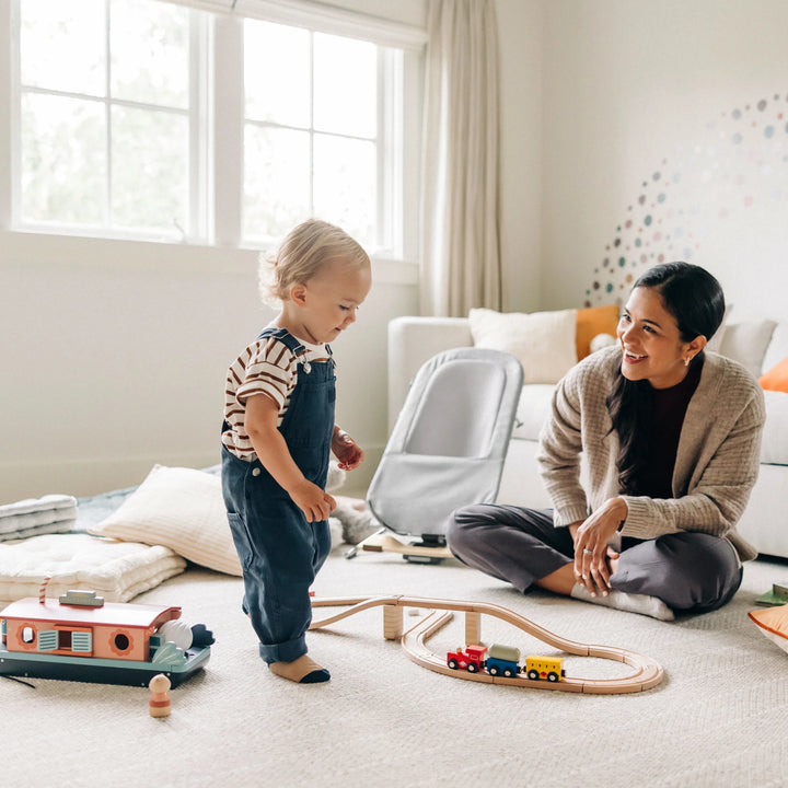 Toddler playing with wooden toys in a bright family living room while the UPPAbaby Mira Bouncer in Stella grey sits nearby, showcasing the modern ergonomic baby bouncer with breathable mesh seat and lightweight wood-accented frame.