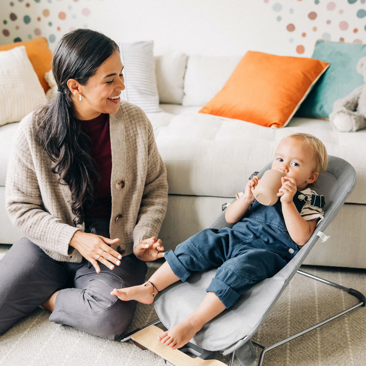 Toddler sitting and drinking from a sippy cup in the UPPAbaby Mira Bouncer in Stella grey while a parent interacts nearby, showcasing the ergonomic toddler-support design, breathable mesh seat, and lightweight modern frame in a bright family living room.