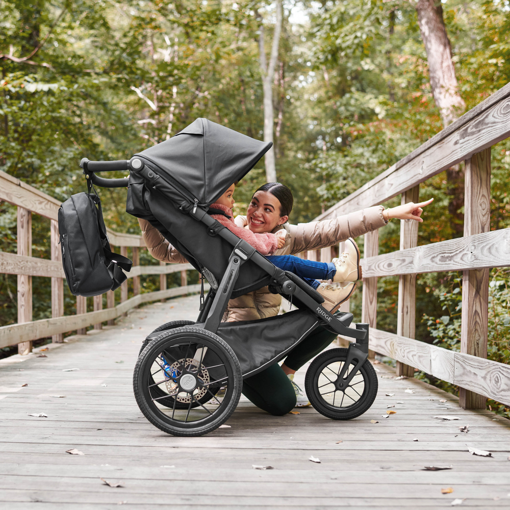 UPPAbaby Ridge active jogging stroller in Jake with a charcoal fabric and carbon frame with foam handlebars. This image shows a mother stopped on the bridge with her little one in the Ridge stroller pointing off the bridge excitedly. The matching changing backpack is attached to the handlebars.