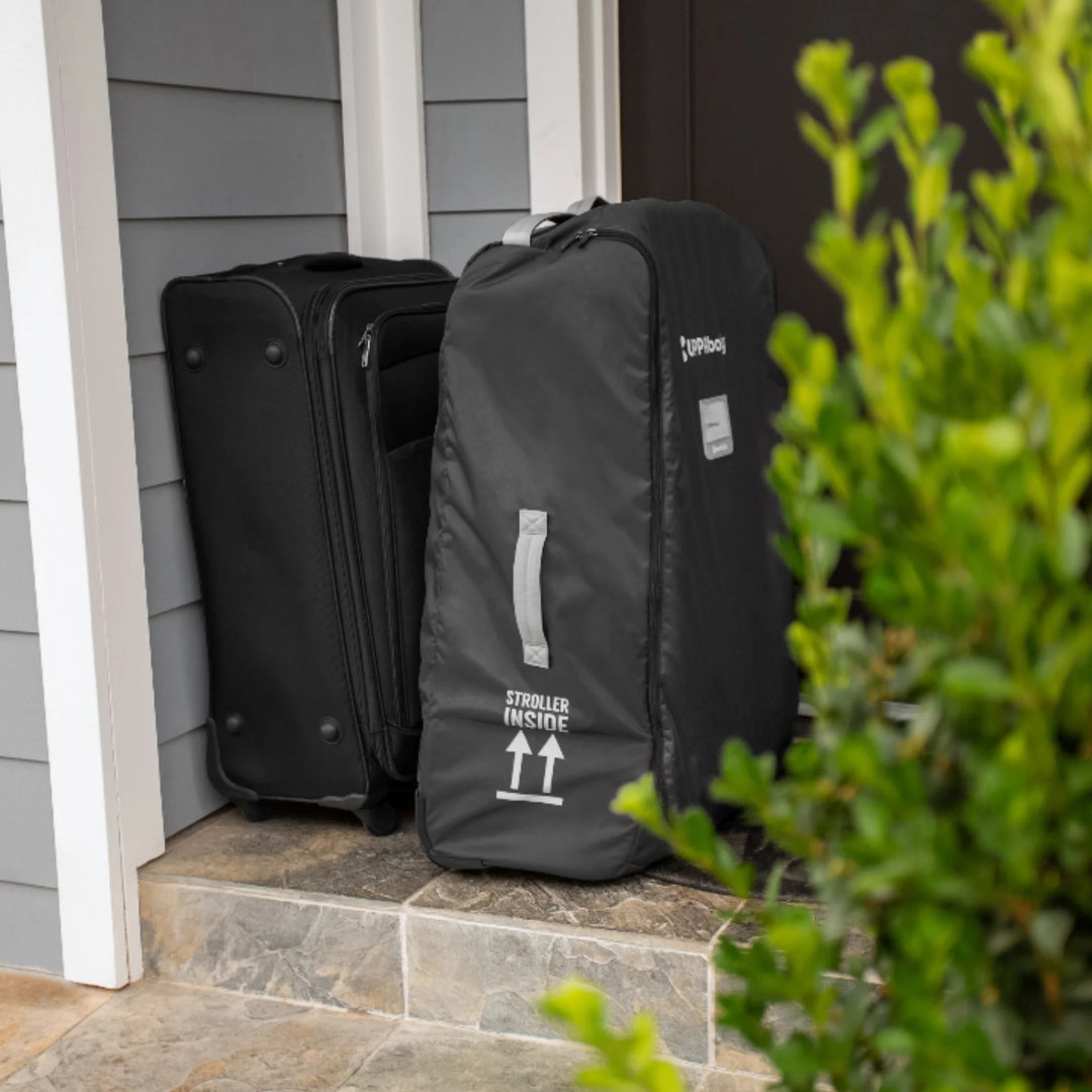 “UPPAbaby VISTA and CRUZ stroller travel bag in black beside a suitcase on a front doorstep.”