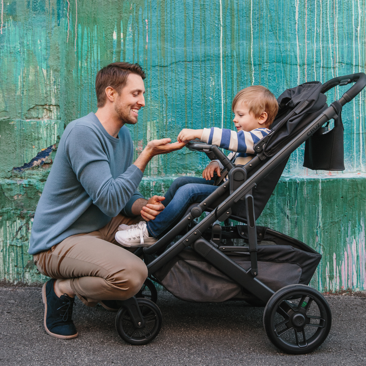 This is a shot of a Dad kneeling down to his child in the stroller who is handing him some snacks from their snack tray on the Cruz V2 stroller in the Jake fashion