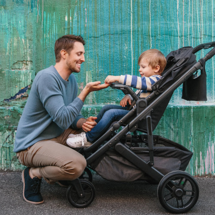 Dad interacting with toddler in UPPAbaby Vista or Cruz stroller fitted with UPPAbaby Snack Tray accessory, outdoors.