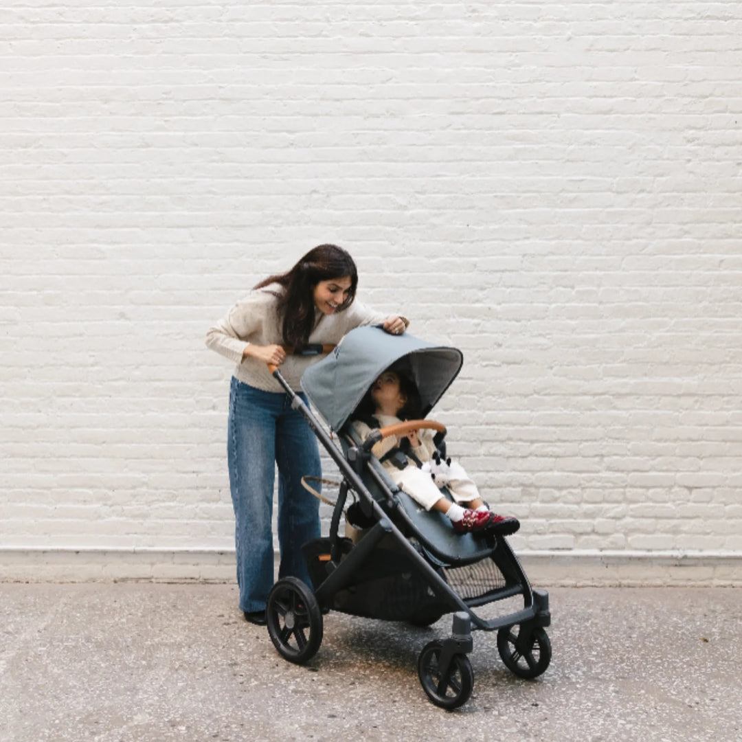 Mother leaning over the UPPAbaby Vista V3 stroller in the Dillian colourway while her toddler sits comfortably in the seat with the sun canopy extended. Lifestyle scene highlighting the Vista V3’s premium leather handlebar, large all-terrain wheels, spacious storage basket and ergonomic toddler seating.