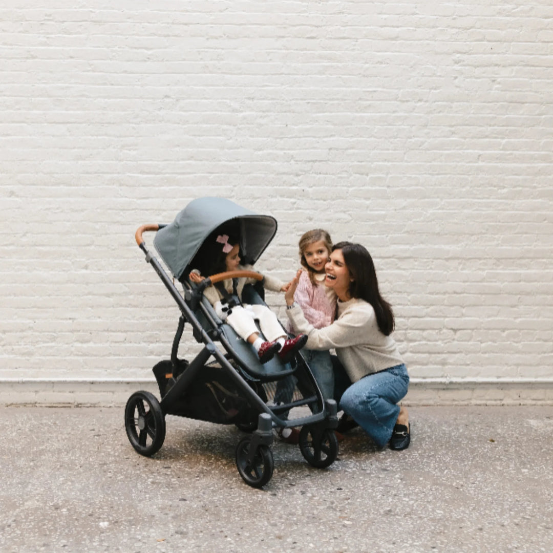 Mother with two young children interacting beside the UPPAbaby Vista V3 stroller in the Dillian colourway, featuring the toddler seat with extended canopy, premium leather handlebar and large all-terrain wheels. Lifestyle family scene showcasing the comfort, durability and everyday usability of the Vista V3 pushchair.
