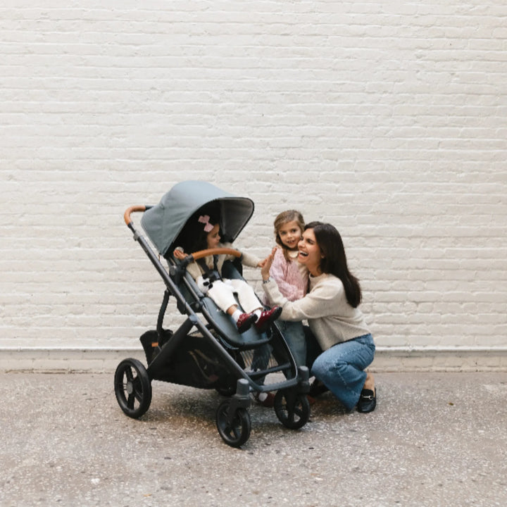 Mother with two young children interacting beside the UPPAbaby Vista V3 stroller in the Dillian colourway, featuring the toddler seat with extended canopy, premium leather handlebar and large all-terrain wheels. Lifestyle family scene showcasing the comfort, durability and everyday usability of the Vista V3 pushchair.