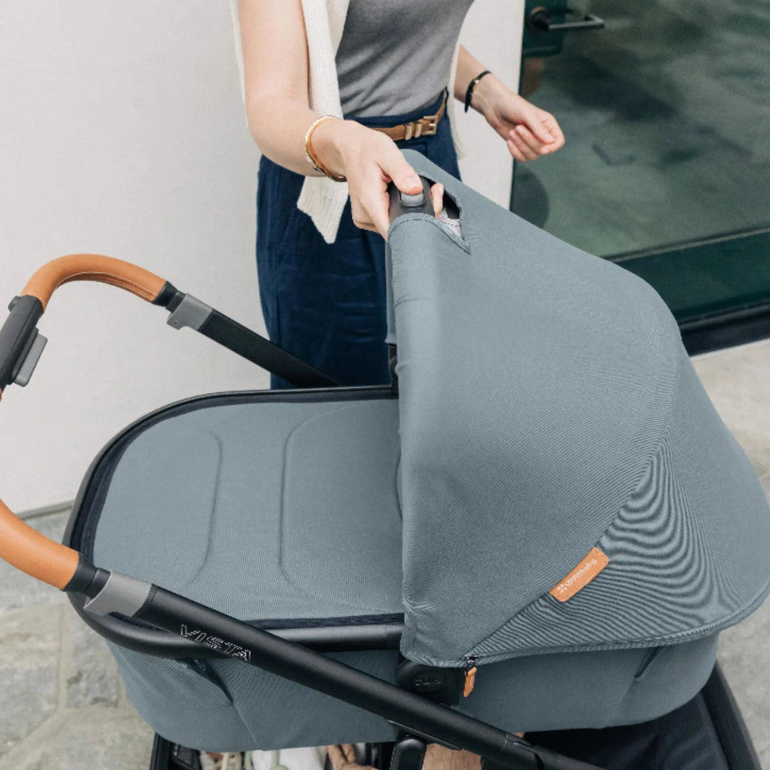 Parent lifting the UPPAbaby Vista V3 carrycot in the Dillian colourway using the easy one-hand release button, showcasing the ergonomic leather handlebar, premium canopy fabric and sturdy Vista frame. Lifestyle image highlighting the convenience, quality and functionality of the Vista V3 newborn carrycot system.