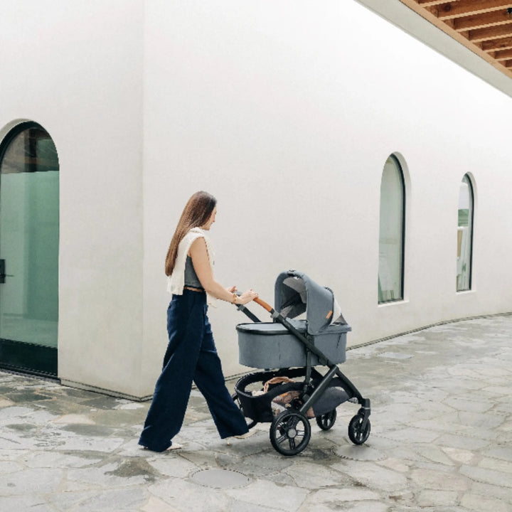 Mother walking outdoors with the UPPAbaby Vista V3 stroller in the Dillian colourway, using the newborn carrycot on the chassis. Lifestyle image showcasing the premium leather handlebar, sleek modern frame design, smooth-rolling all-terrain wheels and spacious carrycot for comfortable newborn travel.