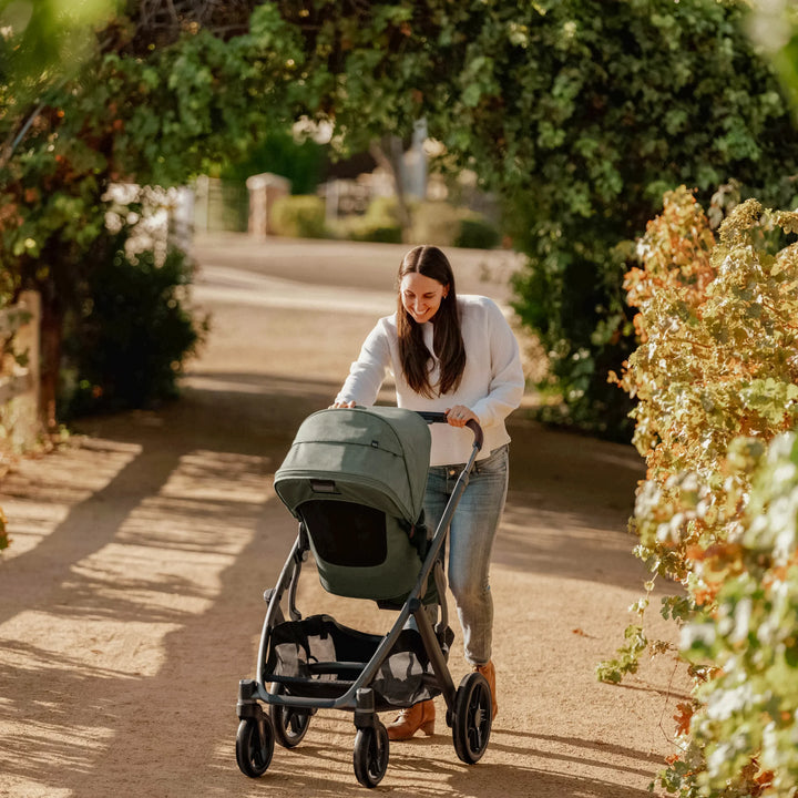Parent pushing the UPPAbaby Vista V3 stroller in the Evelyn green colourway along a sunny outdoor pathway, showcasing the stroller’s stylish design, large canopy, smooth suspension and all-terrain wheels for everyday family use.