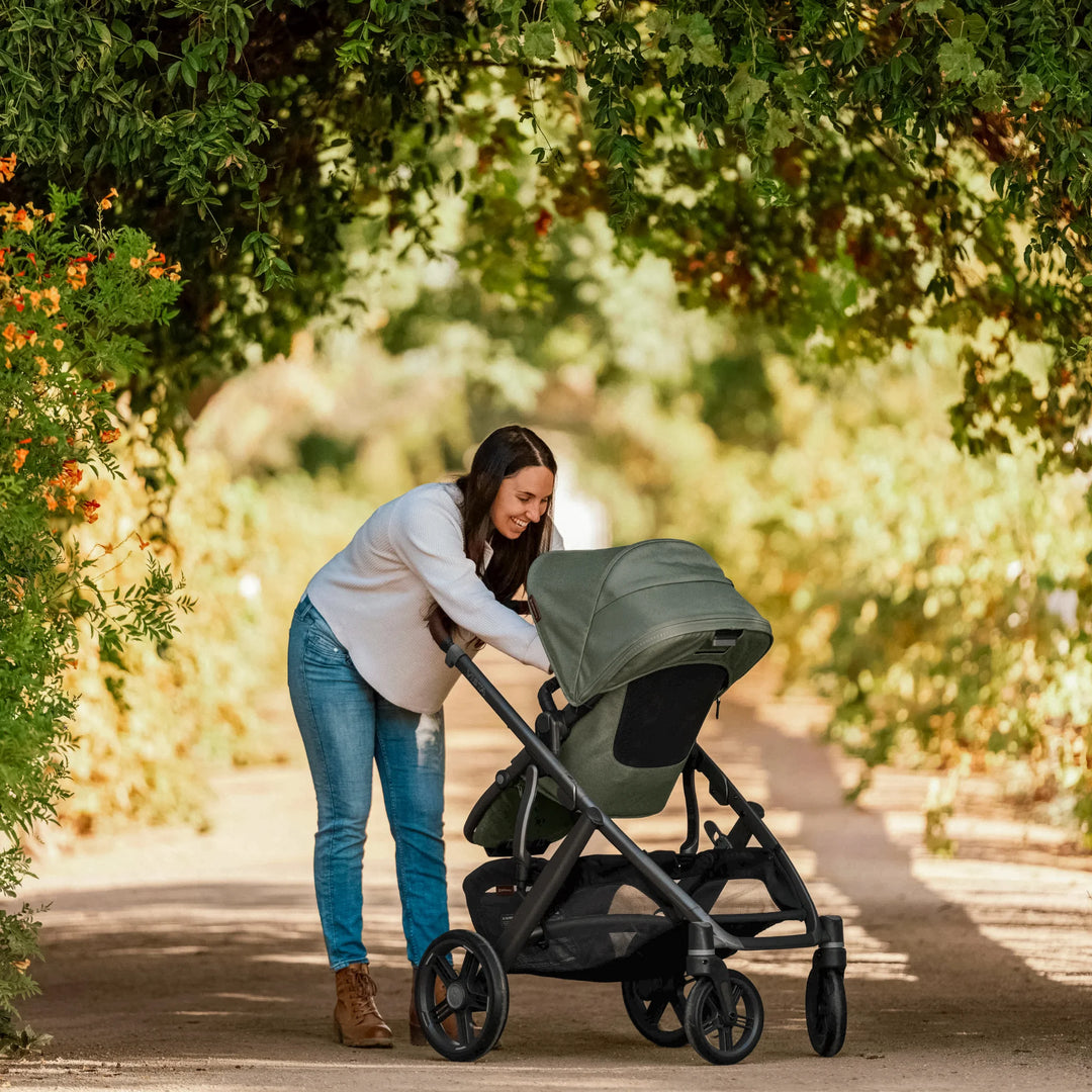 Parent interacting with the UPPAbaby Vista V3 stroller in the Evelyn green colourway on a sunny outdoor path, showcasing the stroller’s ergonomic seat, large canopy, smooth suspension and durable all-terrain wheels.
