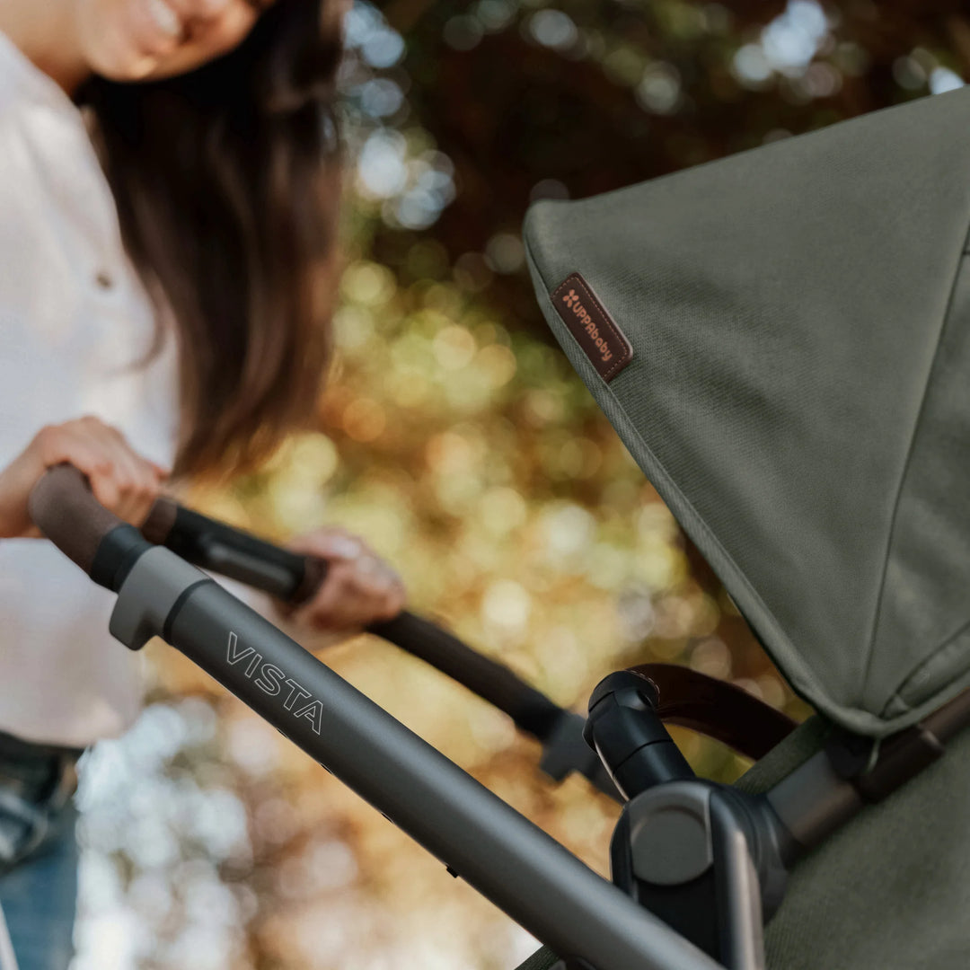Close-up of the UPPAbaby Vista V3 stroller in the Evelyn green colourway being pushed outdoors, showing the premium canopy fabric, leather-accented handlebar, and durable Vista frame branding.