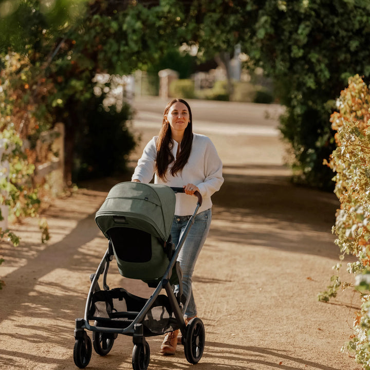 Parent pushing UPPAbaby Vista V3 stroller in the Evelyn green colourway along a sunlit outdoor path, showcasing the stroller’s stylish design, large canopy, smooth all-terrain wheels, and comfortable everyday use.