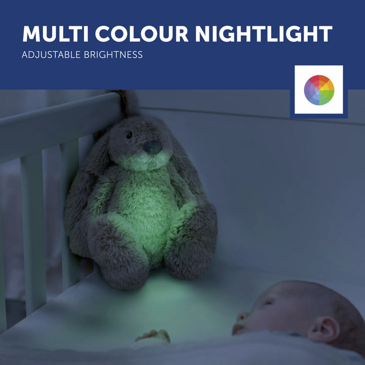 ZAZU Ro the Rabbit Sleep Soother showing multicolour nightlight with adjustable brightness, glowing green in a crib beside a sleeping baby.