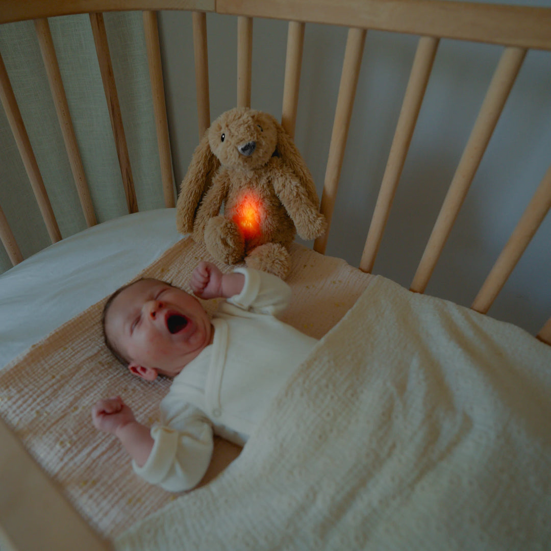 Crying newborn in a cot with the ZAZU Ro the Rabbit Sleep Soother plush night light and sound machine glowing softly to calm and settle baby for sleep.
