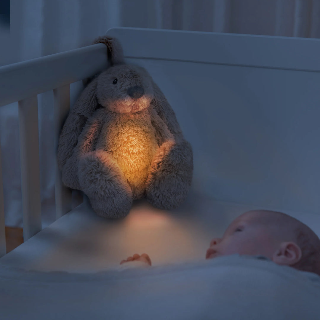 Baby in a cot with the ZAZU Ro the Rabbit Sleep Soother plush night light and sound machine glowing softly, creating a calming bedtime environment for better sleep.