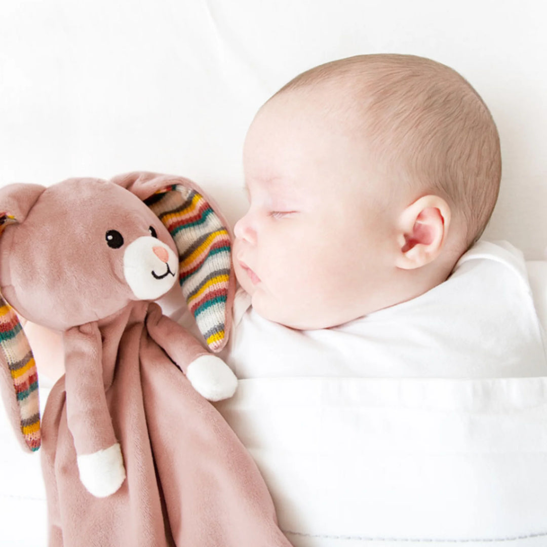 “Sleeping baby cuddling ZAZU Becky bunny comforter with striped ears.”