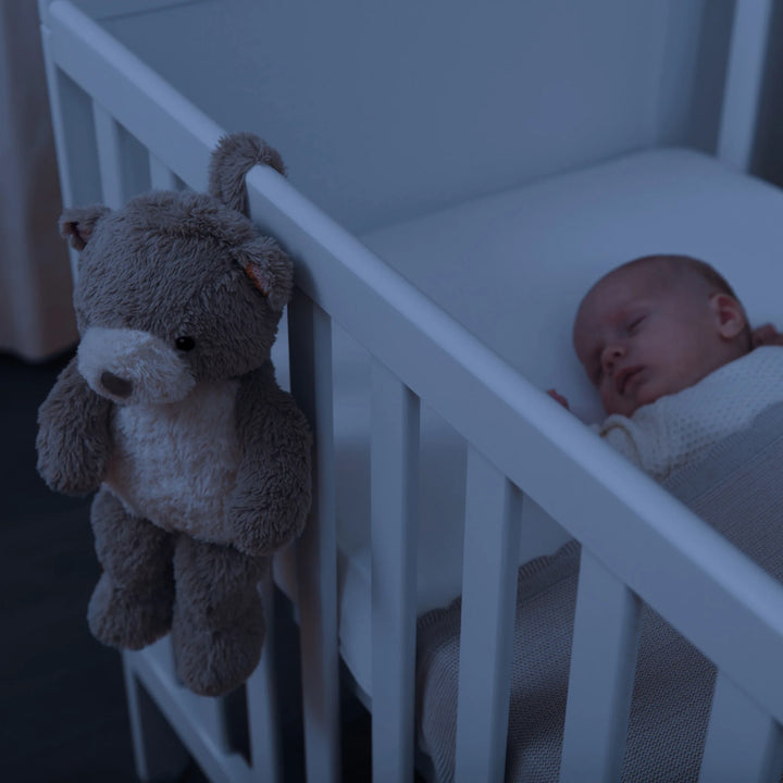 “ZAZU Bruno bear baby soother hanging on a crib at night beside a sleeping infant.”