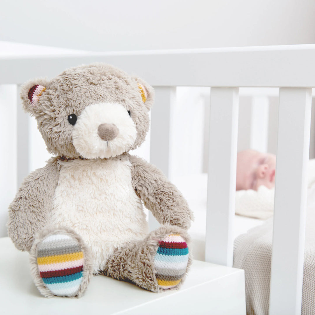 Zazu Bruno Baby Soother – soft grey plush teddy bear with colourful striped paws sitting beside a baby’s cot, perfect comforting sleep companion and nursery decor for newborns.