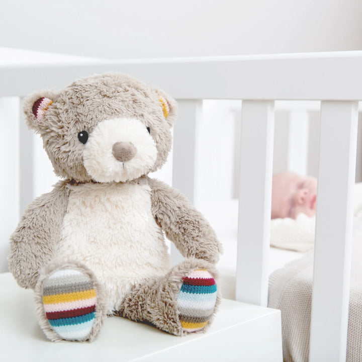 Zazu Bruno Baby Soother – soft grey plush teddy bear with colourful striped paws sitting beside a baby’s cot, perfect comforting sleep companion and nursery decor for newborns.