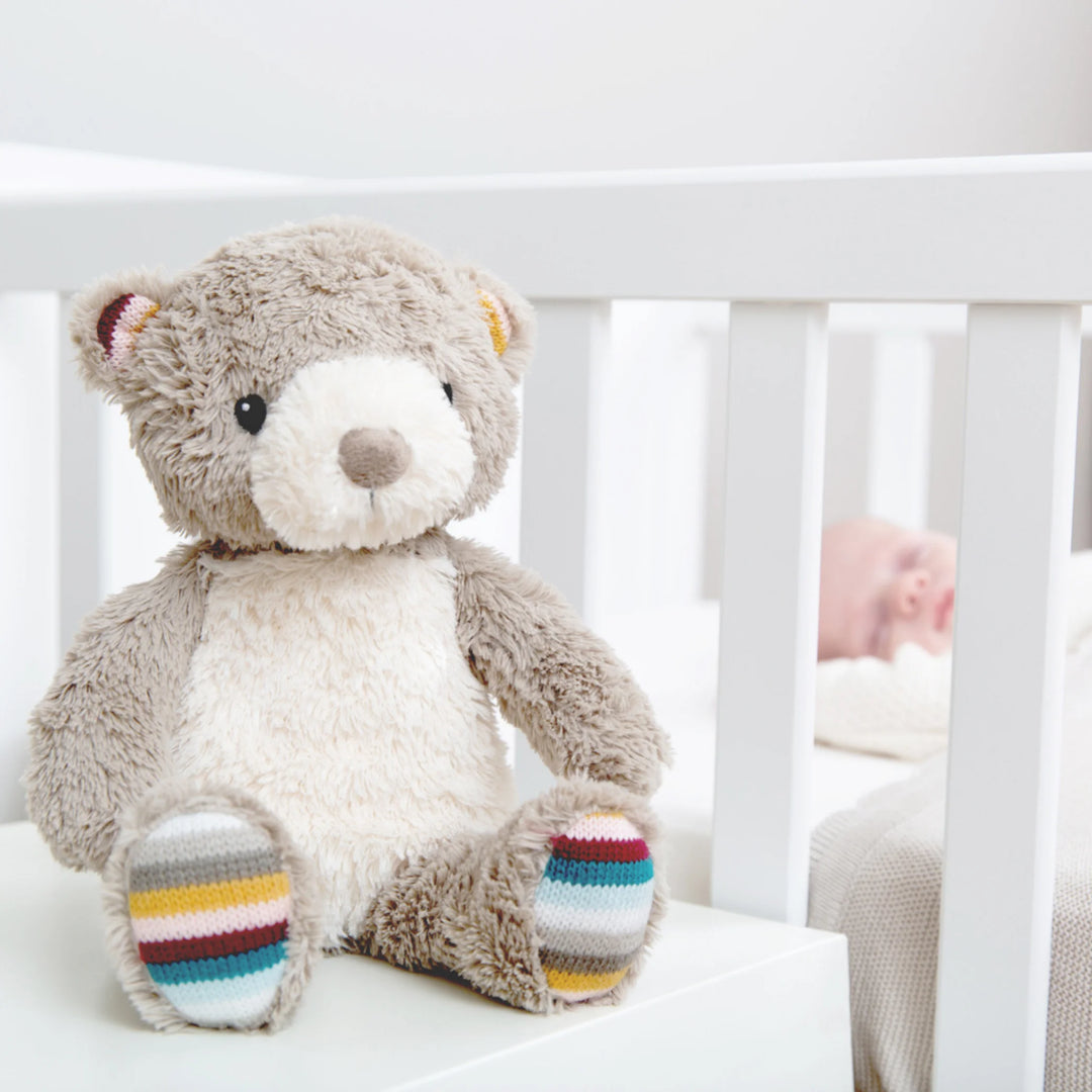 ZAZU Bruno bear baby sleep soother sitting beside a crib with a sleeping newborn in the background.”