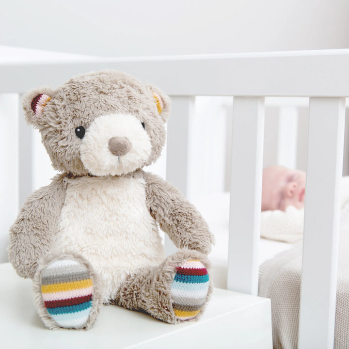 ZAZU Bruno bear baby sleep soother sitting beside a crib with a sleeping newborn in the background.”