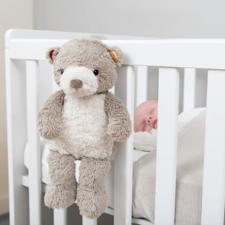 “ZAZU Bruno bear baby soother hanging from a white crib rail beside a sleeping baby.”