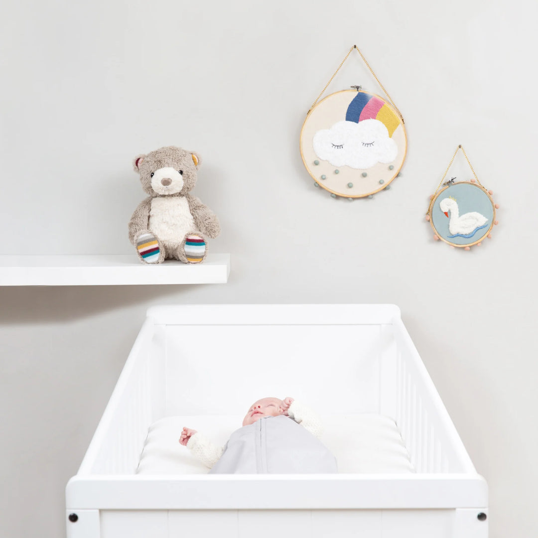 Zazu Bruno Baby Soother plush teddy bear displayed on nursery shelf beside baby sleeping peacefully in white cot, stylish and soothing sleep aid for newborns and nursery decor.
